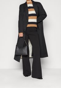 Abrigo largo negro sobre un suéter a rayas en tonos beige, blanco y azul marino, combinado con pantalones negros acampanados y un bolso negro con cadena.