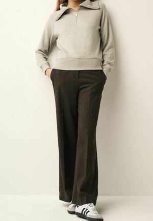 Osoba oblečená v béžovém svetru s krátkým zipem, tmavě hnědých širokých kalhotách a bílých teniskách s černými pruhy stojící před světlou zdí.