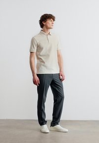 Νεαρός άνδρας που στέκεται, φοράει μπεζ κοντομάνικο πόλο πουκάμισο, σκούρο γκρι παντελόνι και λευκά αθλητικά παπούτσια με φόντο μια απλή επιφάνεια.