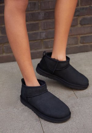 Piernas con botas negras tipo slip-on hasta el tobillo con forro suave, de pie sobre concreto cerca de una pared de ladrillos.