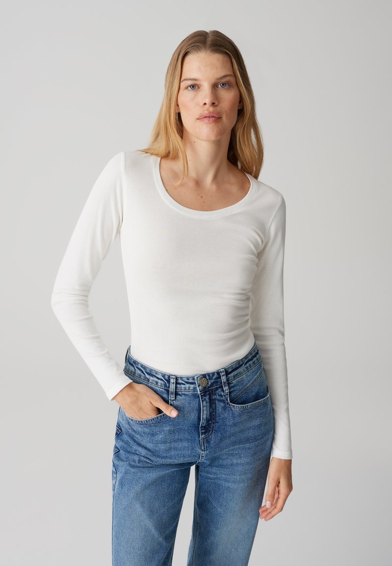 Maglietta bianca a maniche lunghe con scollo rotondo, abbinata a jeans blu a vita alta. Il tessuto sembra liscio e leggermente aderente.