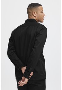 Black long-sleeve shirt with a structured collar, buttoned cuffs, and a smooth texture. The back view shows a simple, minimalist design.