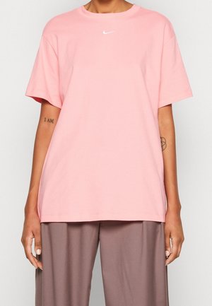 Personne portant un t-shirt Nike ample rose clair et un pantalon marron à larges jambes, avec des tatouages visibles sur les deux avant-bras.