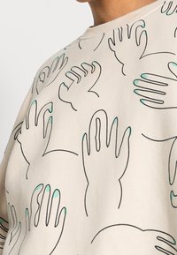 Lichtbeige sweatshirt met een grafisch ontwerp van handen, omlijnd in zwart en voorzien van teal vingertoppen. Zachte stoftextuur.