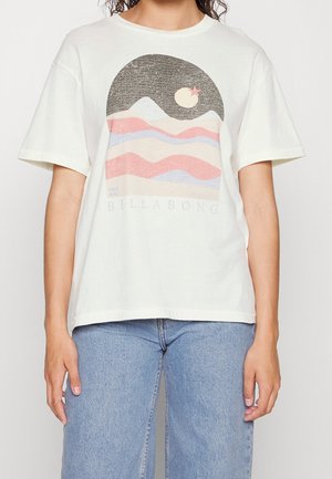 Osoba ubrana w białą koszulkę z pastelowym nadrukiem gór i księżyca oraz napisem "BILLABONG", w zestawie z jasnoniebieskimi jeansami.