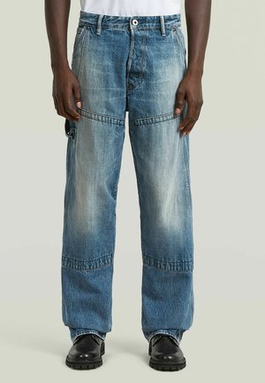 Mann trägt lockere blaue Jeans mit ausgewaschenen Details und schwarzen Stiefeln vor schlichtem Hintergrund.