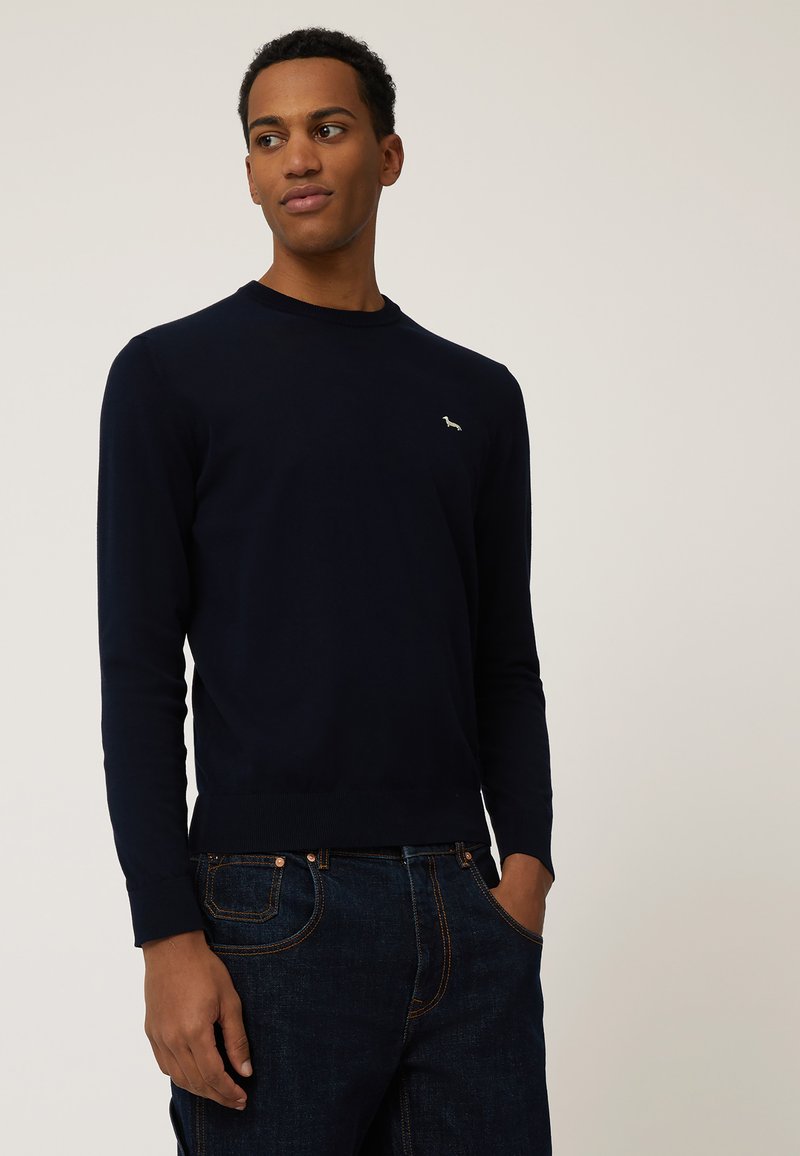 Marineblauer Strickpullover mit langen Ärmeln, geripptem Halsausschnitt und Bündchen, auf der Brust mit einem kleinen gestickten Logo. Getragen mit dunkler Jeans.