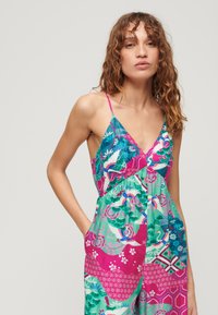 Un mono colorido que presenta una mezcla de patrones florales y geométricos en turquesa, rosa y verde. Incluye tirantes finos y bolsillos laterales.