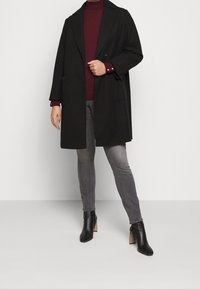 Manteau en laine noire avec un col cranté, une fermeture croisée et des poches latérales, associé à un col roulé bordeaux et un jean gris. Bottines noires à talons.