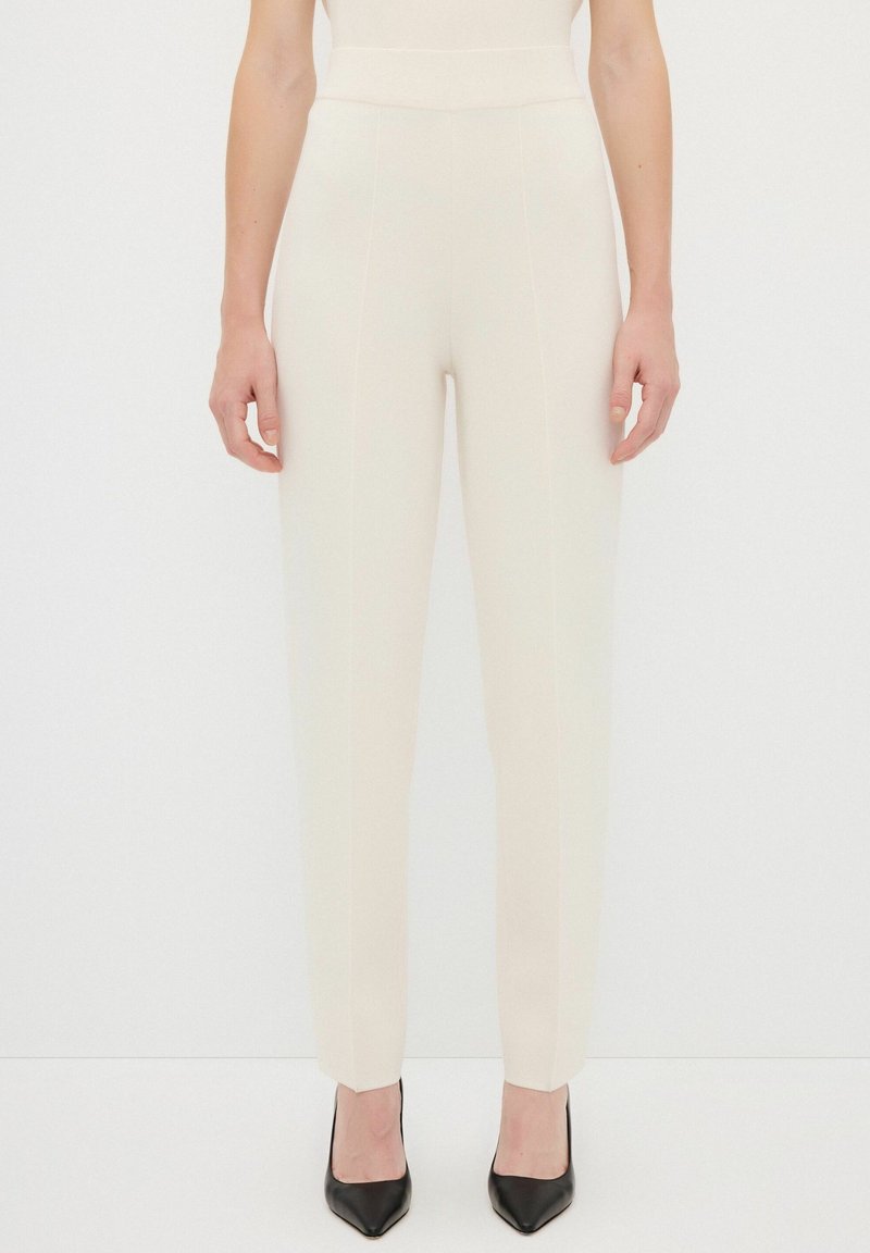 Pantalons sur mesure couleur crème avec une taille haute, une coupe fuselée et des coutures visibles. Associés à des chaussures à talons hauts noires.