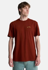 Camiseta de algodón color óxido con escote redondo, mangas cortas y un pequeño logotipo dorado en el pecho. Ajuste casual y textura suave.