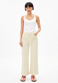 Beige wijde jeans met een hoge taille en zijzakken, gecombineerd met een witte tanktop met subtiele verticale stiksels. Zwarte sandalen met rode accenten.