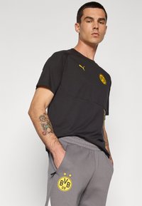 Musta lyhythihainen urheilupaita, jossa on keltainen logo ja akcentit, yhdistettynä harmaisiin jogginghousuihin, joissa on yhteensopiva logo taskussa.