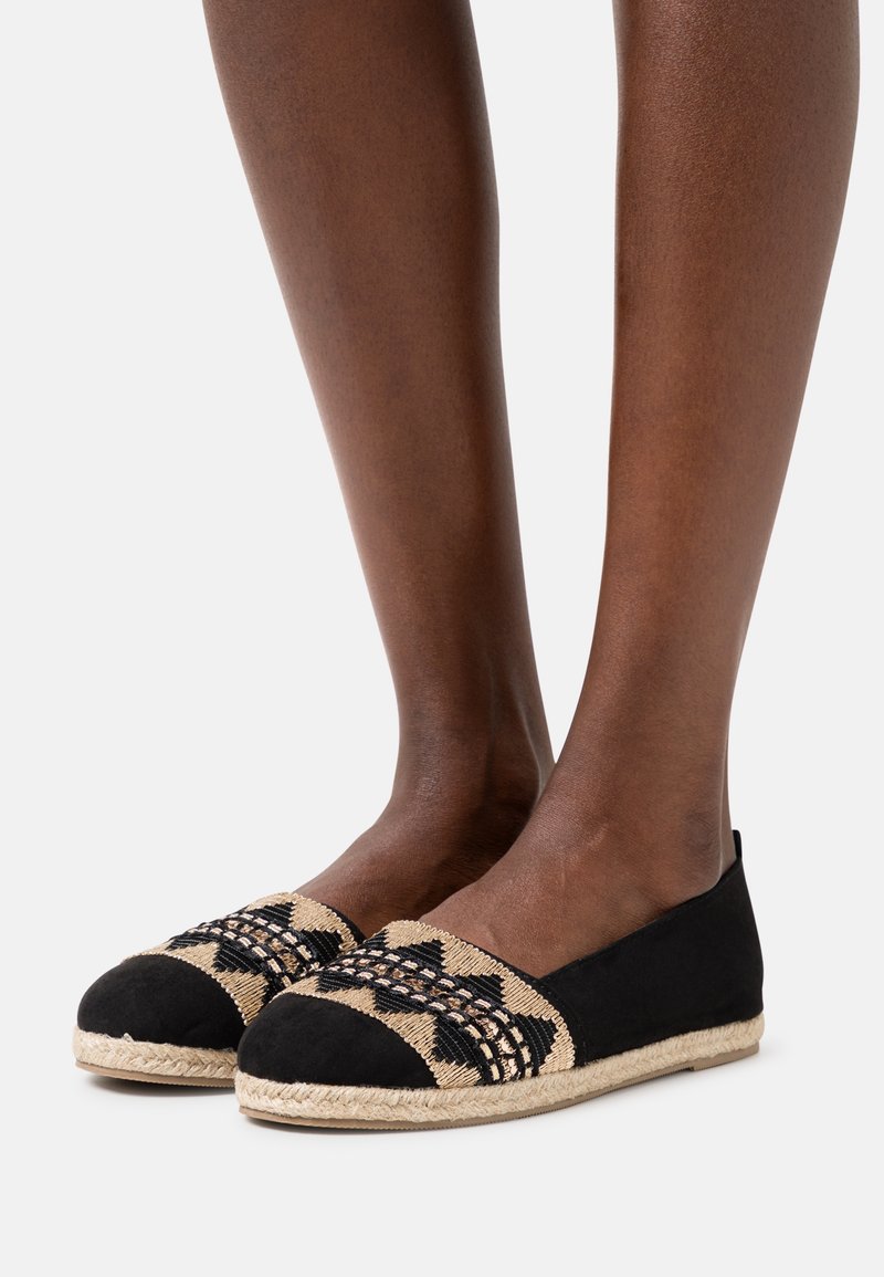 Pieds portant des espadrilles plates tissées noires et beiges avec des détails à motifs et des semelles en corde, présentées sur un fond clair uni.