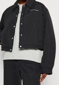 Sort cropped puffer-jakke med en blank finish, trykknapper og logo-detalje. Har en bred krave og let elastiske manchetter.