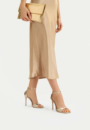 Kobieta w złotej satynowej sukience midi, metalicznych złotych sandałach na wysokim obcasie, trzymająca złotą kopertówkę na jednolitym tle.