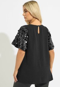 Top nero con maniche corte decorate con paillettes nere, chiusura a occhiello sul retro e corpo in tessuto testurizzato, che offre una vestibilità ampia.