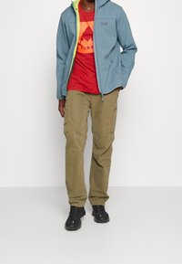 Giacca softshell azzurro chiaro con zip verde neon, indossata sopra una t-shirt grafica rossa. Pantaloni cargo verdi oliva e calzature nere completano il look.