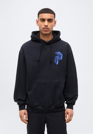 Junger Mann trägt einen schwarzen Hoodie mit blauem geometrischem Muster auf der Brust, steht vor einem schlichten hellgrauen Hintergrund.