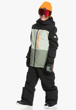 Tenue de ski composée d'une veste isolante verte et noire avec un logo orange, associée à un pantalon noir et un bonnet orange, complétée par des lunettes de ski miroir.