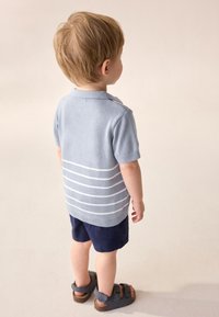 Lyseblå strikket polo med korte ærmer og hvide striber, kombineret med marineblå shorts. Sandaler med justerbare stropper. Billede af barn set bagfra.