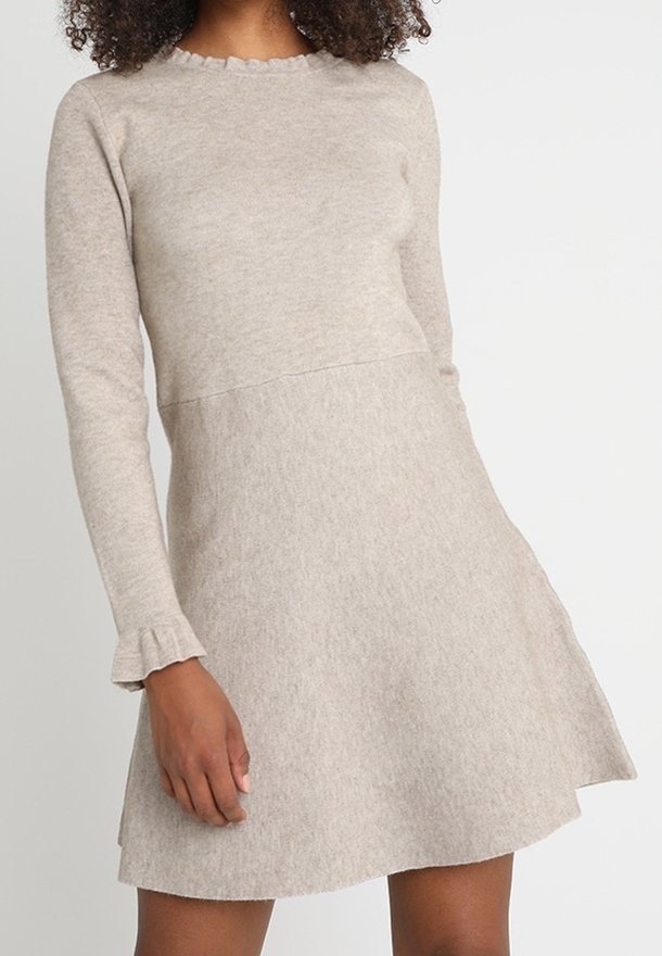Robe en tricot beige à manches longues avec bords festonnés sur l'encolure et les poignets, taille ajustée et jupe trapèze portée par une personne debout.