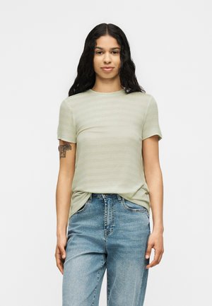 Jeune personne avec de longs cheveux bouclés portant une chemise à manches courtes vert clair rentrée dans un jean bleu taille haute, debout devant un fond uni.