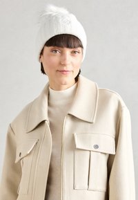 Gorro de punto blanco con un pompón esponjoso en la parte superior y logotipo bordado. Combinado con una chaqueta cremosa con cremallera que cuenta con dos bolsillos.