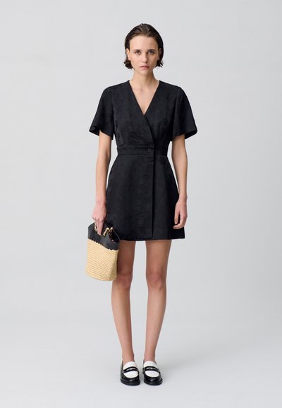 Vestito nero avvolgente con maniche corte e una texture floreale, abbinato a una borsa intrecciata in paglia e mocassini in bianco e nero.