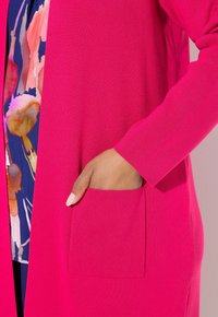 Cardigan fuchsia avec texture côtelée, à l'avant ouvert et avec poche latérale. Porté par-dessus un haut à motifs floraux aux couleurs vives.