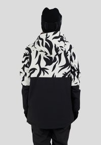 Persona che indossa una giacca a cappuccio con motivo in bianco e nero, parte inferiore nera uniforme, pantaloni neri, guanti e un berretto nero, di spalle.