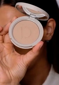 Beige kompaktt puder med präglad "D"-logotyp, förpackad i en cirkulär, ljusgrå plastlåda med ett genomskinligt lock. En hand som håller produkten är synlig.