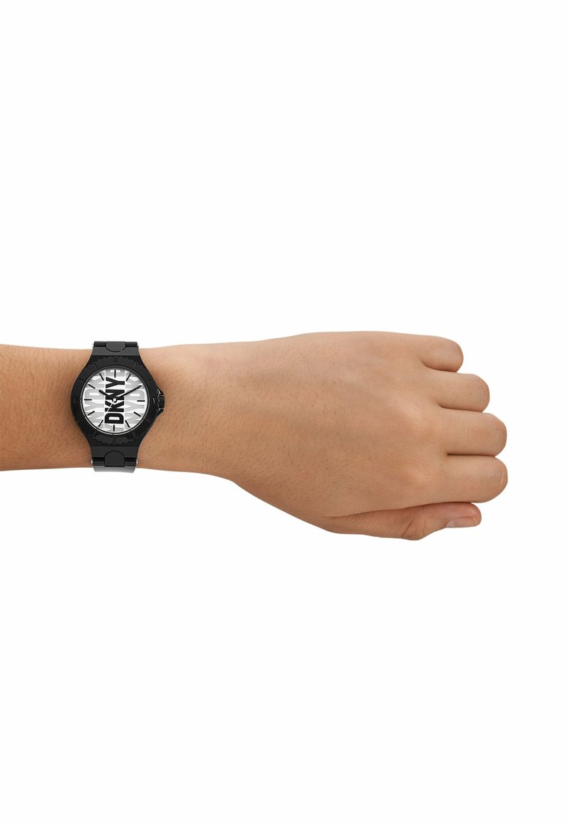Poignet gauche portant une montre DKNY noire avec un cadran blanc et un logo noir en gras, tenue devant un fond blanc uni.