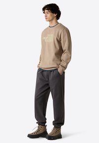 Beige sweatshirt med tekstureret overflade og logo, parret med løstsiddende mørkegrå bukser og brune vandrestøvler med sorte såler og snørebånd.