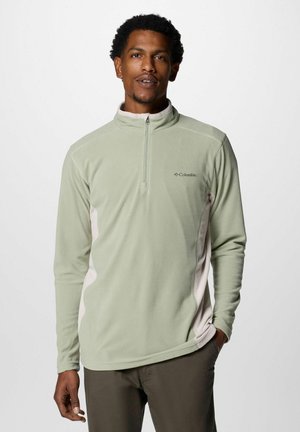 Uomo che indossa un pullover in pile Columbia verde chiaro con zip fino a un quarto e pannelli laterali beige, in piedi con una mano in tasca.