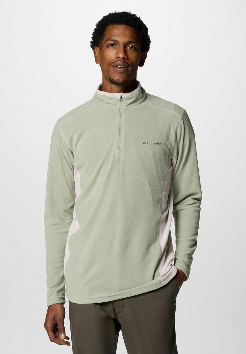 Uomo che indossa un pullover in pile Columbia verde chiaro con zip fino a un quarto e pannelli laterali beige, in piedi con una mano in tasca.