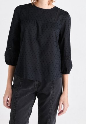 Kobieta w czarnej, fakturowanej bluzce z bufkami o długości do łokcia i czarnych, wysokich spodniach dżinsowych na jednolitym białym tle.