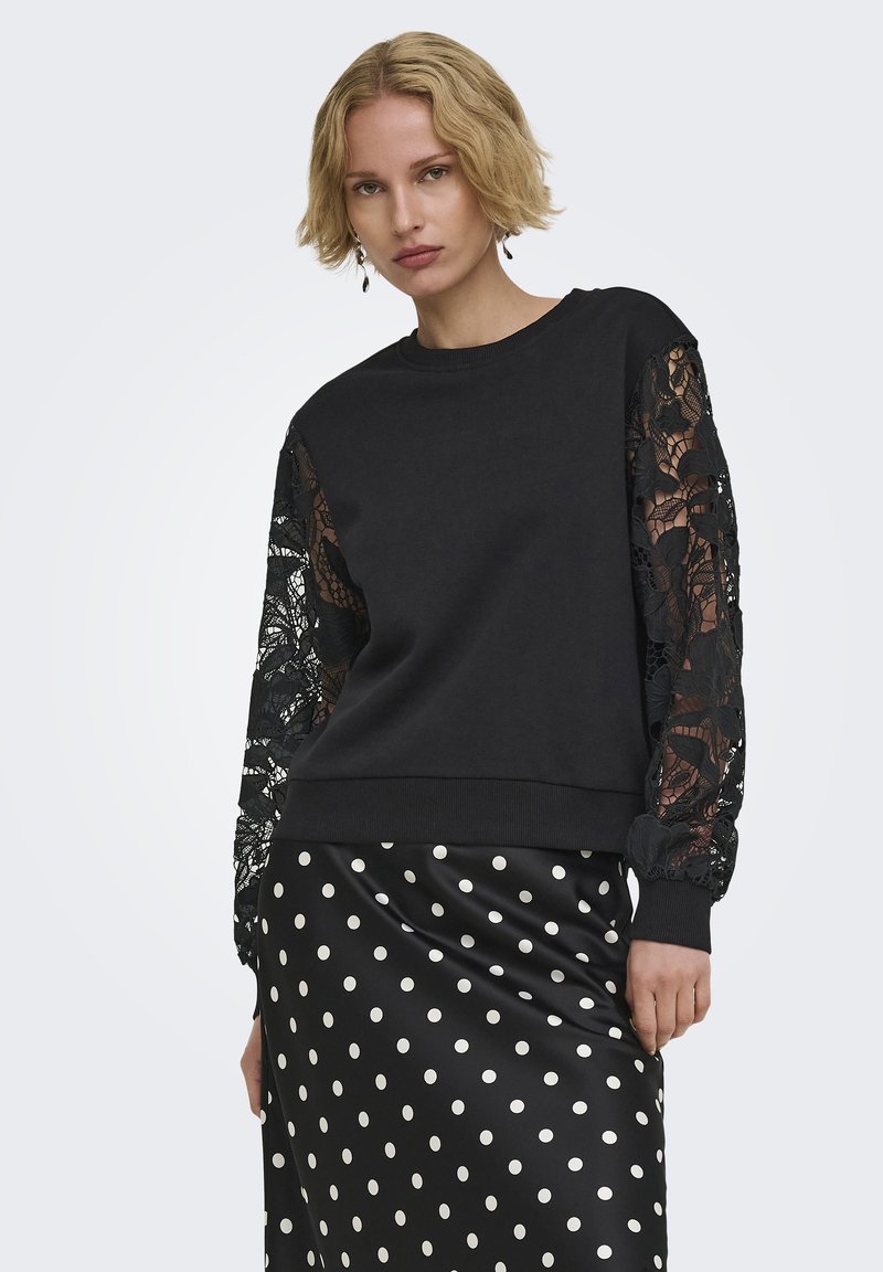 Femme blonde porte un pull noir avec des manches en dentelle florale et une jupe en satin noire à pois blancs sur un fond uni.