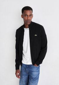 Μαύρο μπουφάν με φερμουάρ και ριχτές μανσέτες, συνδυασμένο με λευκό μπλουζάκι με στρογγυλή λαιμόκοψη που έχει πράσινες λεπτομέρειες και μπλε τζιν παντελόνι. Το λογότυπο Lacoste εμφανές.