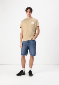 Tan short-sleeve t-shirt with a front pocket design, paired with blue denim shorts and black sneakers with white socks.