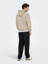 Sweat à capuche beige avec une coupe décontractée et une capuche ajustable, porté avec un pantalon noir. Tissu léger, design minimaliste, et ourlet côtelé.
