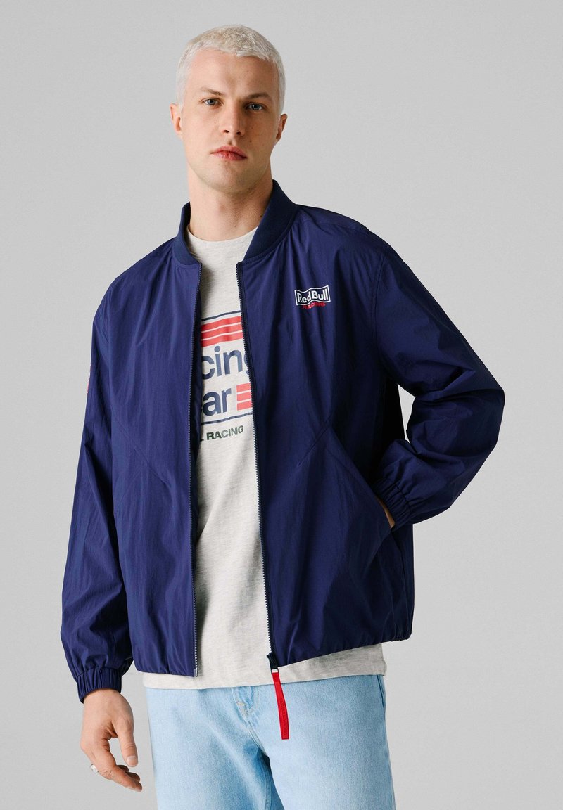 Blonde man die een marineblauwe Red Bull Racing-jas draagt over een lichtgekleurde grafische T-shirt en lichtblauwe spijkerbroek, staand tegen een effen grijze achtergrond.