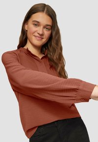 Ruda bluzka o teksturowym wykończeniu, z długimi rękawami, zapinana na guziki z falbaną przy kołnierzu, noszona na czarnych spodniach.