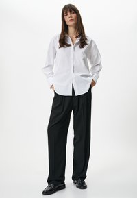 Camicia bianca con bottoni e maniche lunghe, abbinata a pantaloni neri a gamba larga. Il modello sta in piedi con le mani nelle tasche, indossando scarpe nere.