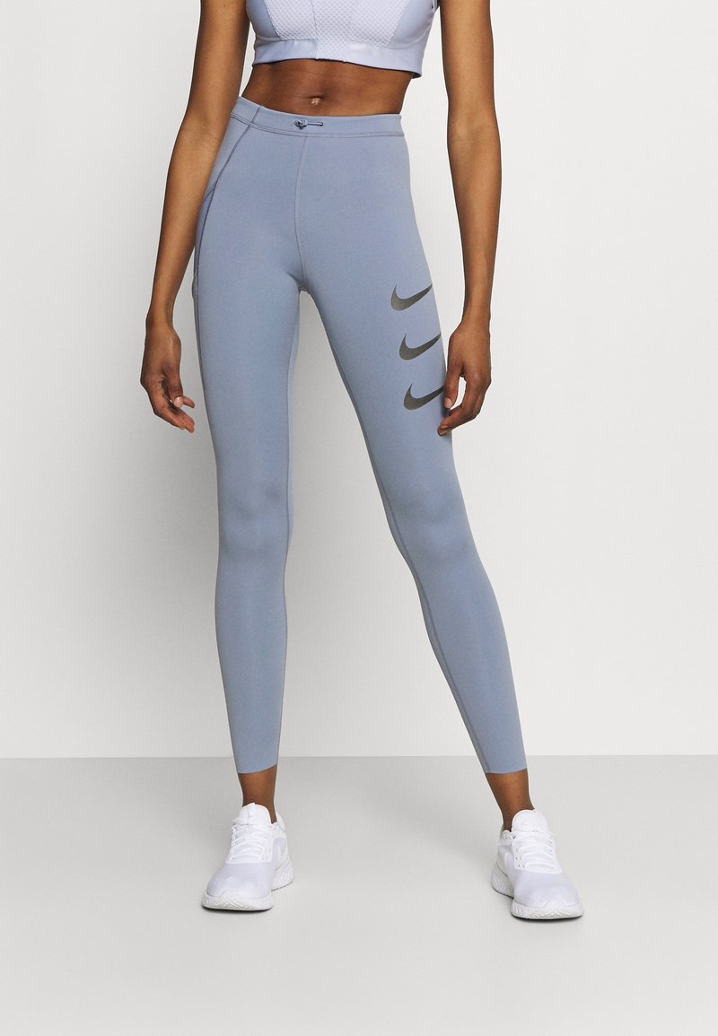 Leggings bleu clair avec une taille haute, texture lisse, et trois swooshes gris foncé sur la cuisse gauche. Associés à des chaussures de sport blanches.