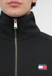 Black zip-up jacket with a high collar, featuring a silver zipper and a small blue, red, and white "TOMMY JEANS" logo on the chest.