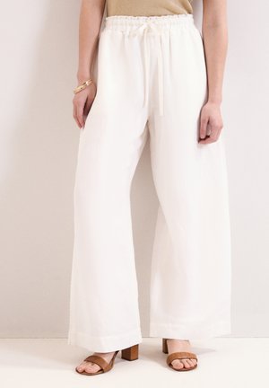 Persoon die losse witte brede broek met elastische tailleband en bruine sandalen met hakken draagt, staand tegen een effen achtergrond.