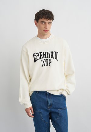 Joven con cabello rizado que lleva un suéter blanco Carhartt WIP y vaqueros azules, de pie frente a un fondo liso y claro.