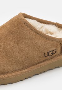 Pantofola in suede marrone con una morbida fodera in pelliccia, suola in gomma texturizzata e logo visibile sul lato. Design classico slip-on.