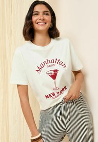 Biela bavlnená tričko s červeným potlačou, ktoré zobrazuje pohár martini, nápis "Manhattan Est1870" a "New York Social Club". Krátke rukávy, voľný strih.